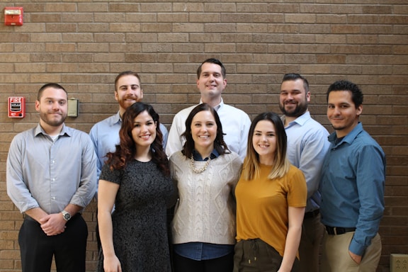A group of eight people are standing in front of a brick wall, smiling at the camera. They are casually dressed and positioned closely together, suggesting a friendly and professional atmosphere. A fire alarm is visible on the wall to the left.