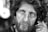 The black and white photograph captures the close-up of a middle-aged man with a rugged appearance. His curly hair is disheveled, and he has a thick beard. His eyes stare intently, conveying deep emotion. The skin shows visible signs of weathering and life experience.