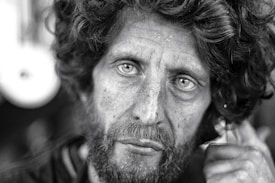 The black and white photograph captures the close-up of a middle-aged man with a rugged appearance. His curly hair is disheveled, and he has a thick beard. His eyes stare intently, conveying deep emotion. The skin shows visible signs of weathering and life experience.