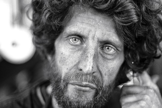 The black and white photograph captures the close-up of a middle-aged man with a rugged appearance. His curly hair is disheveled, and he has a thick beard. His eyes stare intently, conveying deep emotion. The skin shows visible signs of weathering and life experience.