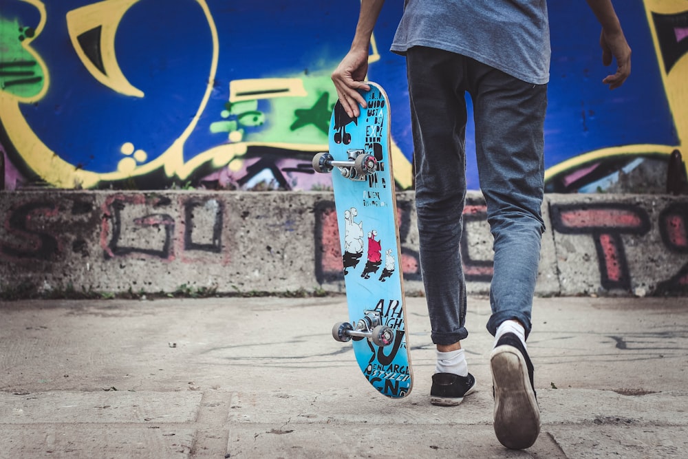 Persona sosteniendo patineta azul caminando cerca de graffiti