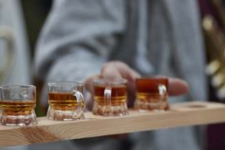 A person holds a wooden tray with four small glass cups filled with a golden-brown liquid. The cups resemble miniature beer mugs, and the focus is on the liquid shining under light.