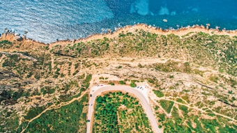 An aerial view captures a rugged coastline with clear blue ocean waters meeting a rocky shore. The landscape has a variety of vegetation and winding pathways. A road curves along the bottom of the image, with several parked cars visible.