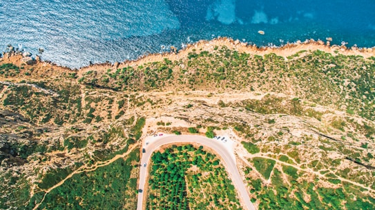 An aerial view captures a rugged coastline with clear blue ocean waters meeting a rocky shore. The landscape has a variety of vegetation and winding pathways. A road curves along the bottom of the image, with several parked cars visible.