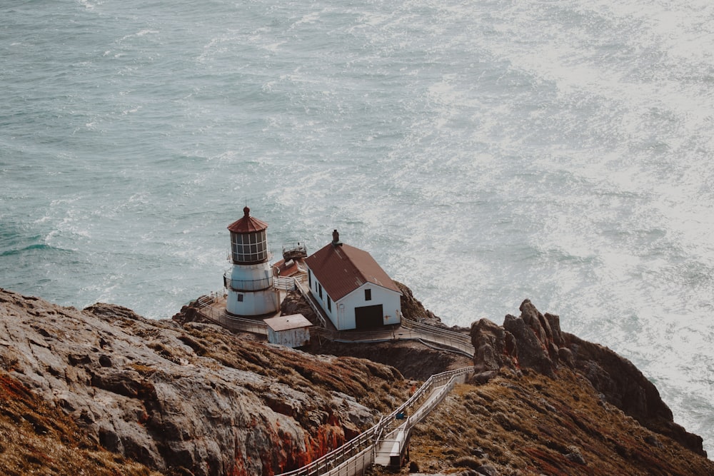 Luftaufnahme eines weißen und braunen Betonhauses neben einem Leuchtturm auf einer grauen Bergklippe in der Nähe eines Gewässers bei Tag