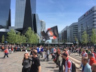A bustling urban scene with modern glass skyscrapers in the background and a crowd of people gathered in the foreground. A variety of individuals, many wearing red and white sports jerseys, are present along with an individual waving a large flag. The weather appears sunny, and the trees lining the street are lush and green.