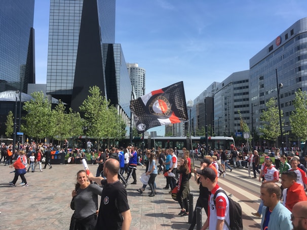 A bustling urban scene with modern glass skyscrapers in the background and a crowd of people gathered in the foreground. A variety of individuals, many wearing red and white sports jerseys, are present along with an individual waving a large flag. The weather appears sunny, and the trees lining the street are lush and green.