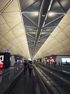 A spacious airport terminal with a high, geometric ceiling that features triangular patterns. People are walking along a moving walkway, and several shops with bright signs are visible on the sides. The atmosphere is modern and bustling.