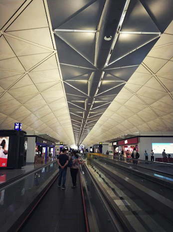 A spacious airport terminal with a high, geometric ceiling that features triangular patterns. People are walking along a moving walkway, and several shops with bright signs are visible on the sides. The atmosphere is modern and bustling.