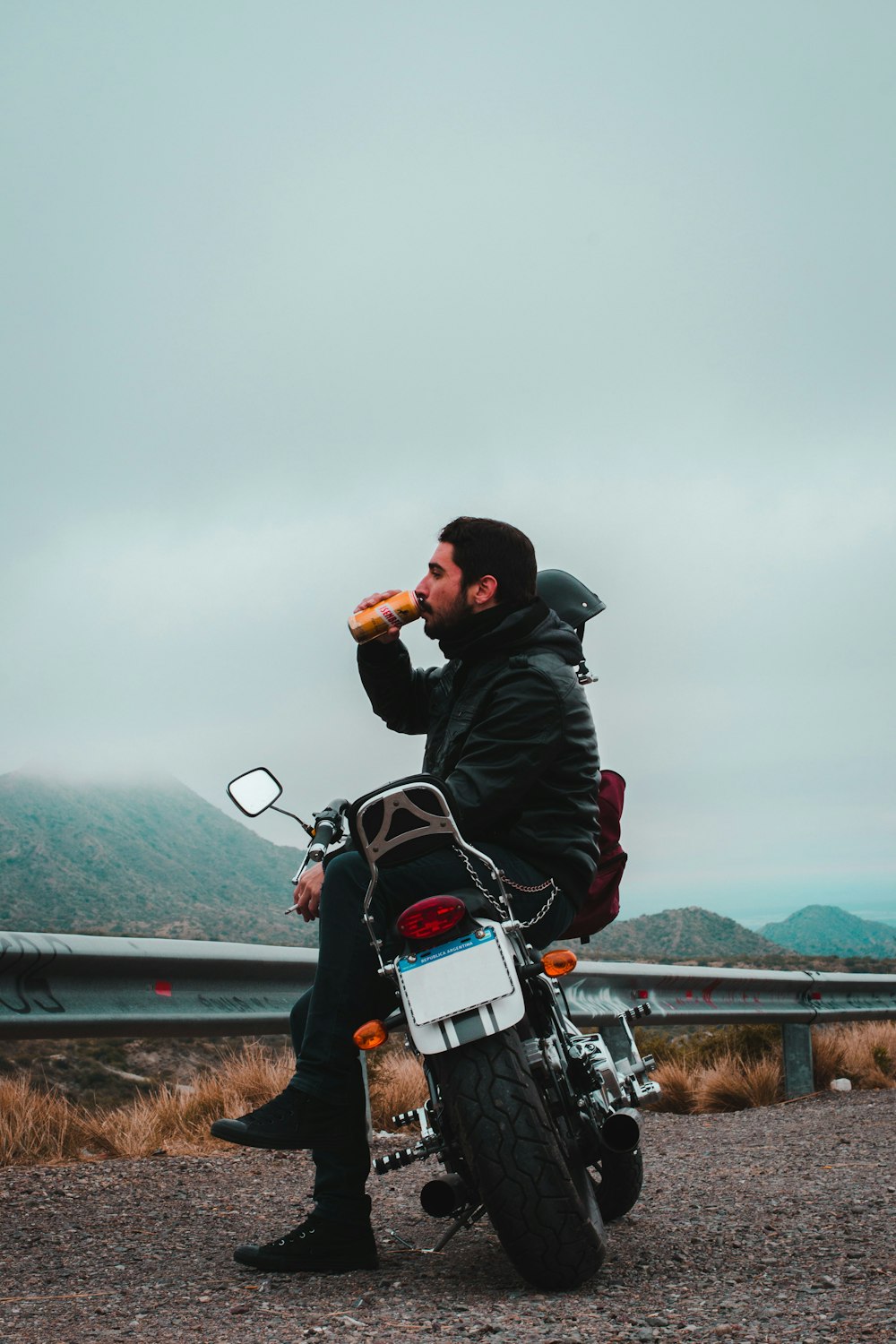 昼間、暗い曇り空の下、山を見下ろす缶で飲み物を飲みながらバイクに座る男