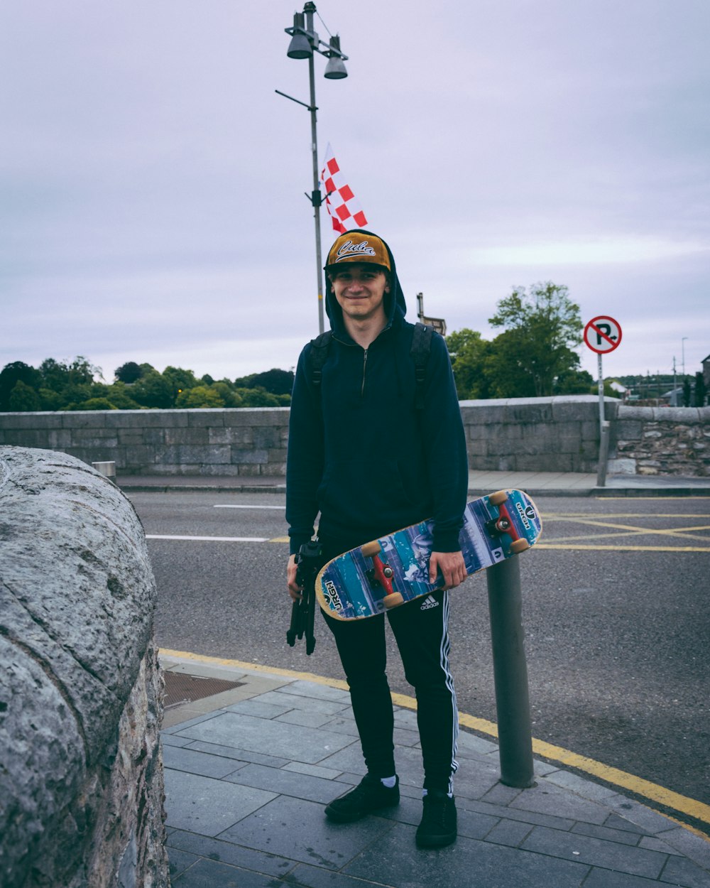 smiling man wearing black hoodie holding skateboard and standing beside bollard across street