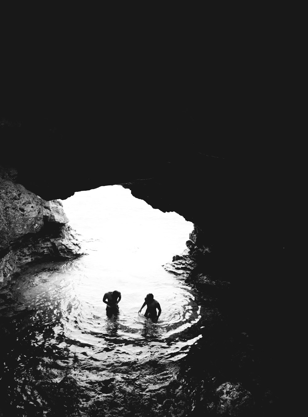 grayscale photo of two men in cave with body of water