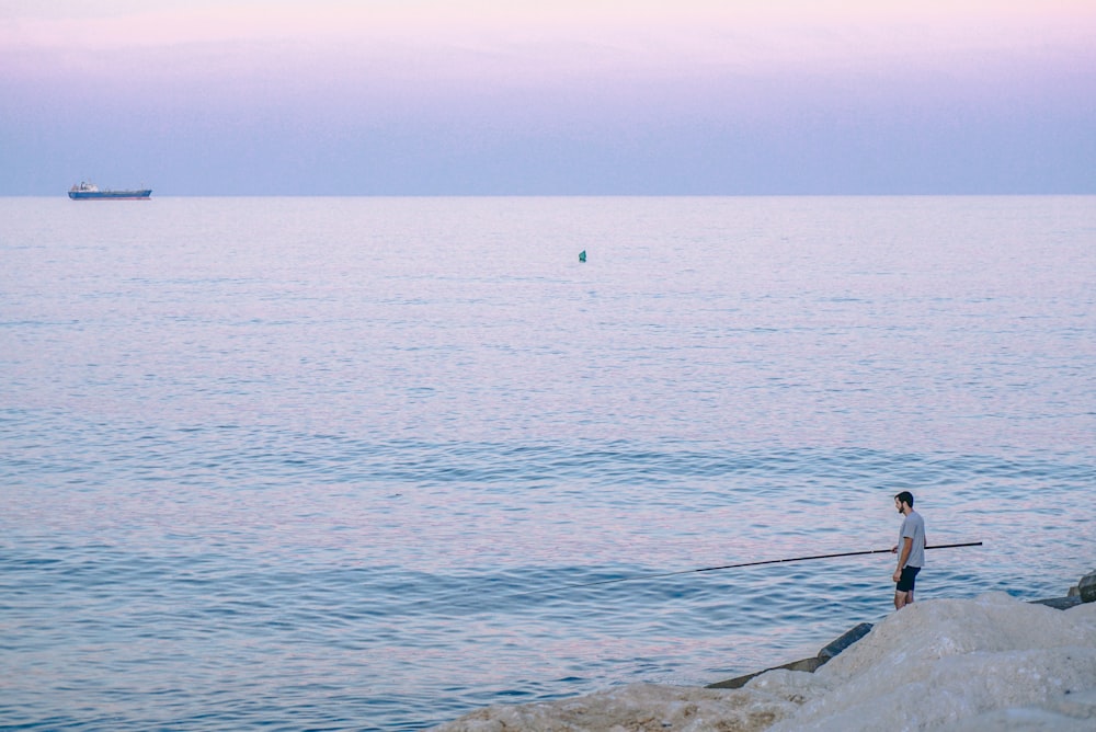 person standing on rock holding stick facing body of water during golden hour