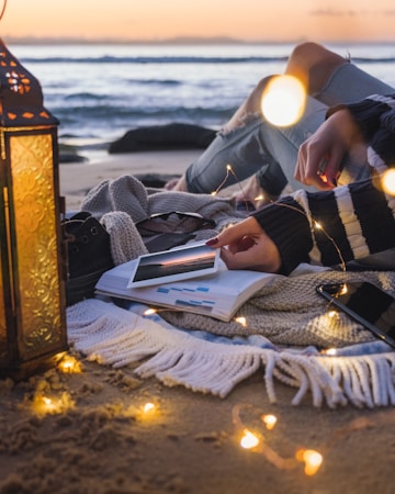 A cozy shoreline setup featuring a person sitting on a blanket. The person holds a photo while resting near an open book with highlighted sections. Warm string lights surround the area, adding a soft glow. An ornate lantern is positioned nearby, enhancing the tranquil ambiance. The sea is visible in the background, with a warm sunset sky.