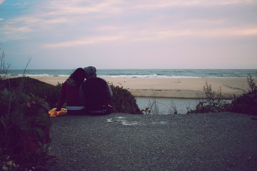 man and woman sits on gray asphalt way over ocean under cloudy sky during daytime