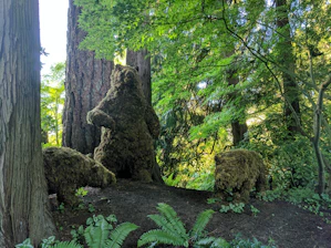 Three bear sculptures made from foliage and moss are set within a lush green forest. One bear is standing upright, while the other two are positioned on all fours. The surrounding area has tall trees, ferns, and other greenery, creating a natural and tranquil environment.