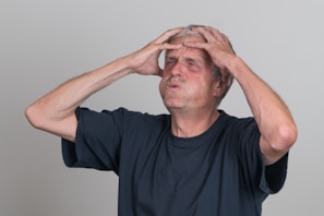 An older man with gray hair and a beard stands against a plain backdrop. He appears to be expressing frustration or stress, as he is holding his head with both hands, and his facial expression is tense. He is wearing a dark-colored t-shirt.