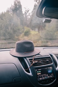 A broad-brimmed hat rests on the dashboard of a car. The car's media console displays a radio station, and the windshield offers a view of a blurred, serene outdoor landscape with trees visible through it.