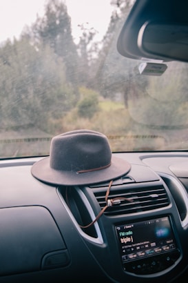 A broad-brimmed hat rests on the dashboard of a car. The car's media console displays a radio station, and the windshield offers a view of a blurred, serene outdoor landscape with trees visible through it.
