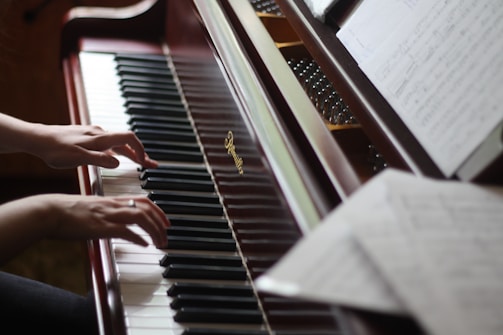A pair of hands gracefully playing the keys of an upright piano. Sheet music is visible on the music stand, and the polished wooden finish of the piano adds a classic touch.