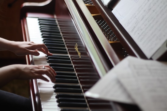 A pair of hands gracefully playing the keys of an upright piano. Sheet music is visible on the music stand, and the polished wooden finish of the piano adds a classic touch.