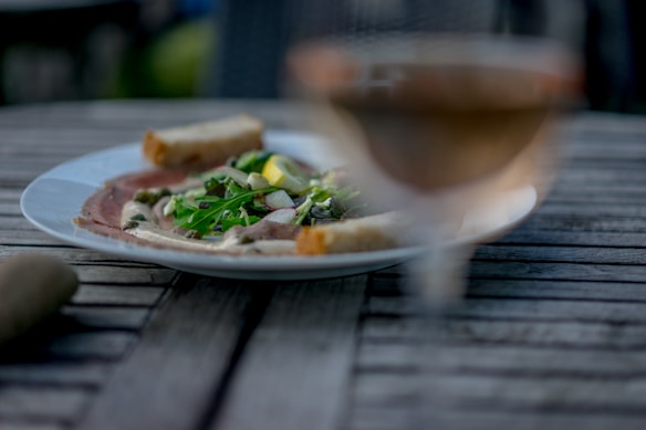 A wooden table hosts a plate of delicately presented food, including leafy greens, a lemon wedge, and a drizzle of dressing. In the foreground, the soft focus includes a glass, possibly containing a beverage, adding an aesthetic blur to the scene.