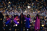 A reflective space filled with numerous small, glowing lights creating an immersive and infinite effect. Several people are engaging with the exhibit, some taking photos or observing quietly. The lights vary in color and intensity, providing a dreamy and ethereal atmosphere.