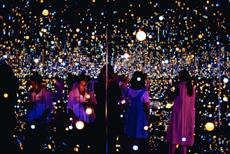 A reflective space filled with numerous small, glowing lights creating an immersive and infinite effect. Several people are engaging with the exhibit, some taking photos or observing quietly. The lights vary in color and intensity, providing a dreamy and ethereal atmosphere.