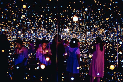 A reflective space filled with numerous small, glowing lights creating an immersive and infinite effect. Several people are engaging with the exhibit, some taking photos or observing quietly. The lights vary in color and intensity, providing a dreamy and ethereal atmosphere.