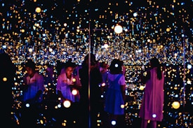 A reflective space filled with numerous small, glowing lights creating an immersive and infinite effect. Several people are engaging with the exhibit, some taking photos or observing quietly. The lights vary in color and intensity, providing a dreamy and ethereal atmosphere.