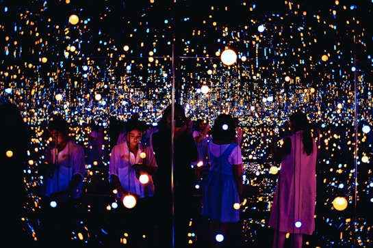 A reflective space filled with numerous small, glowing lights creating an immersive and infinite effect. Several people are engaging with the exhibit, some taking photos or observing quietly. The lights vary in color and intensity, providing a dreamy and ethereal atmosphere.