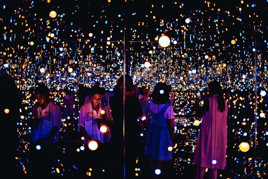 A reflective space filled with numerous small, glowing lights creating an immersive and infinite effect. Several people are engaging with the exhibit, some taking photos or observing quietly. The lights vary in color and intensity, providing a dreamy and ethereal atmosphere.