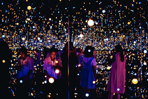 A reflective space filled with numerous small, glowing lights creating an immersive and infinite effect. Several people are engaging with the exhibit, some taking photos or observing quietly. The lights vary in color and intensity, providing a dreamy and ethereal atmosphere.