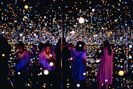 A reflective space filled with numerous small, glowing lights creating an immersive and infinite effect. Several people are engaging with the exhibit, some taking photos or observing quietly. The lights vary in color and intensity, providing a dreamy and ethereal atmosphere.