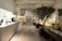 A modern, elegant hotel lobby with a minimalist design features a long reception counter on the left. Several people are working behind the counter. There are large potted plants, contemporary art sculptures, and soft lighting from stylish lamps scattered throughout the space. Comfortable seating areas with beige cushions are arranged on the right side.