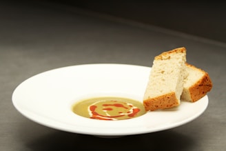 A white plate containing cream soup garnished with a red sauce swirl, accompanied by two slices of bread placed on the edge of the plate. The background is a neutral color, highlighting the simplicity and elegance of the dish.