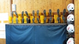 A row of yellow boots is displayed upside down on a shelf, with the soles facing outward. Nearby, several white helmets are stacked vertically on hooks. The backdrop features wooden paneling and blue fabric, likely a part of a maritime or safety equipment storage area.
