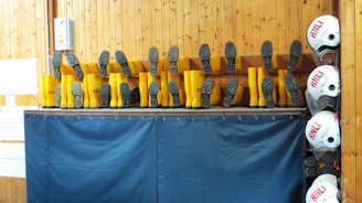 A row of yellow boots is displayed upside down on a shelf, with the soles facing outward. Nearby, several white helmets are stacked vertically on hooks. The backdrop features wooden paneling and blue fabric, likely a part of a maritime or safety equipment storage area.