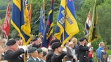 A group of individuals in formal attire and berets stands in formation, holding colorful flags representing various organizations. The scene takes place outdoors, with lush green trees in the background, suggesting a ceremonial or commemorative event.