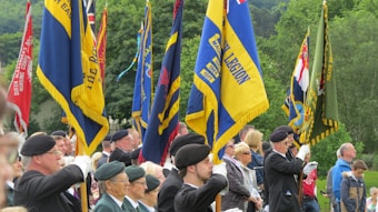 A group of individuals in formal attire and berets stands in formation, holding colorful flags representing various organizations. The scene takes place outdoors, with lush green trees in the background, suggesting a ceremonial or commemorative event.