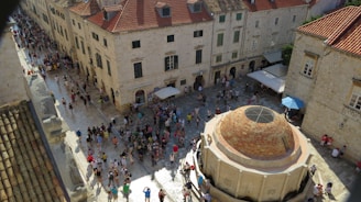 A busy piazza surrounded by old stone buildings with red-tiled roofs. People are gathered around a large circular stone fountain located centrally in the square. A few people are sitting or standing under umbrellas that provide shade in the area.