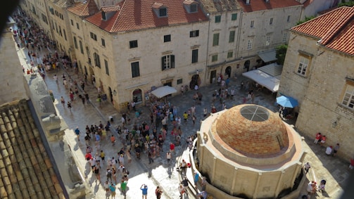 A busy piazza surrounded by old stone buildings with red-tiled roofs. People are gathered around a large circular stone fountain located centrally in the square. A few people are sitting or standing under umbrellas that provide shade in the area.