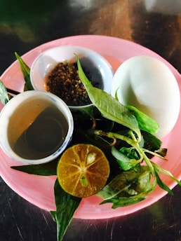 A pink plate holds a small bowl of ground spices, a hard-boiled egg, a cut lime showing its juicy pulp, and fresh green herbs. Next to the bowl is a small cup with a light brown liquid.