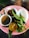 A pink plate holds a small bowl of ground spices, a hard-boiled egg, a cut lime showing its juicy pulp, and fresh green herbs. Next to the bowl is a small cup with a light brown liquid.
