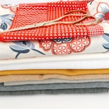 A stack of colorful fabric pieces in various designs and patterns, with a pair of golden scissors placed on top. The fabrics include some with geometric and floral patterns and are arranged neatly in layers.