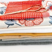 A stack of colorful fabric pieces in various designs and patterns, with a pair of golden scissors placed on top. The fabrics include some with geometric and floral patterns and are arranged neatly in layers.
