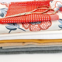 A stack of colorful fabric pieces in various designs and patterns, with a pair of golden scissors placed on top. The fabrics include some with geometric and floral patterns and are arranged neatly in layers.