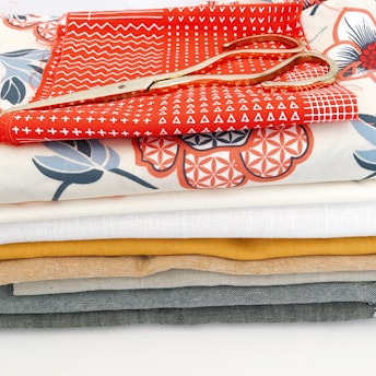 A stack of colorful fabric pieces in various designs and patterns, with a pair of golden scissors placed on top. The fabrics include some with geometric and floral patterns and are arranged neatly in layers.