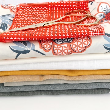 A stack of colorful fabric pieces in various designs and patterns, with a pair of golden scissors placed on top. The fabrics include some with geometric and floral patterns and are arranged neatly in layers.