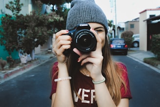 woman wearing red t-shirt and gray knitted cap standing on concrete road using Canon bridge camera during daytime